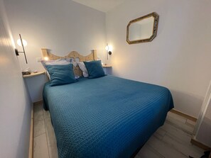 A bedroom with a bed, two bedside tables, a nightstand, a wall-mounted mirror, and a wall socket.