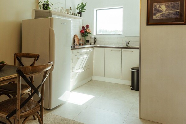Una cocina con armarios blancos, un refrigerador, una ventana y un cubo de basura.