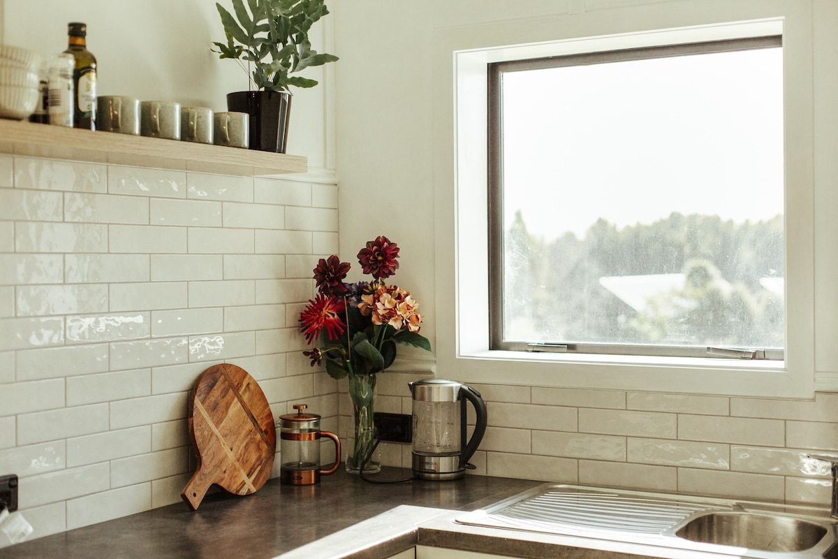 Una cocina con ventana, tabla de madera para cortar, un florero con flores y una tetera.