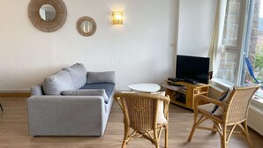 Een woonkamer met een grijze bank, twee rieten stoelen, een klein witte tafel, een televisie op een houten dressoir, en een wandlamp.