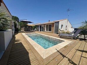 A rectangular swimming pool with a wooden deck, a covered seating area, and a house with a tiled roof.