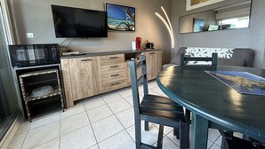 A modern kitchen with a flat-screen TV, microwave, and wooden cabinets. There is a dining table with chairs and a sofa in the living area.