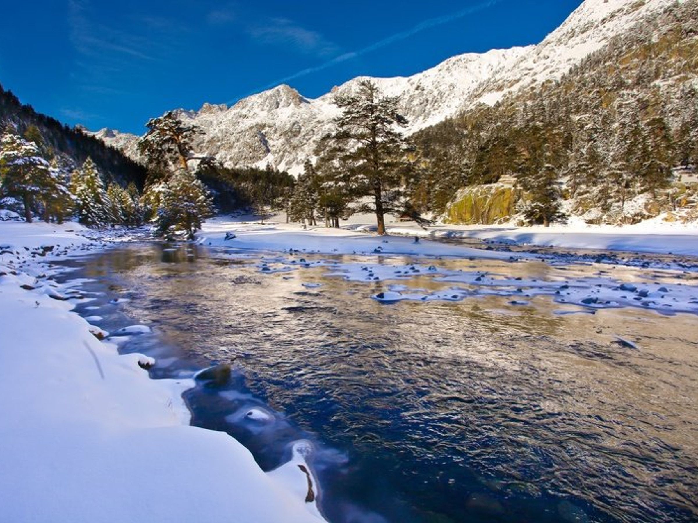 A snow-covered landscape with a river, trees, and mountains.