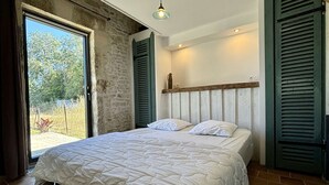 Une chambre à coucher avec un lit, une tête de lit en bois, une fenêtre avec des volets et une vue sur la verdure à l’extérieur.