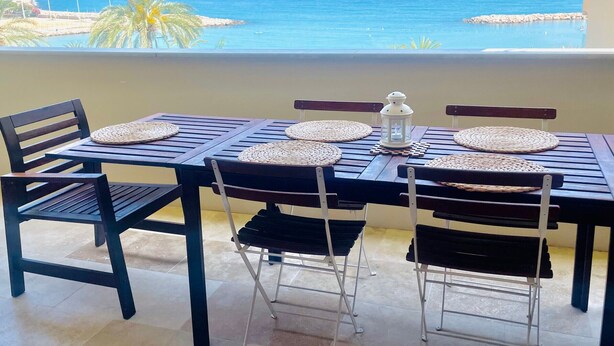 A dining table with woven placemats and a white lantern, set on a balcony with a view of the sea and a distant mountain.