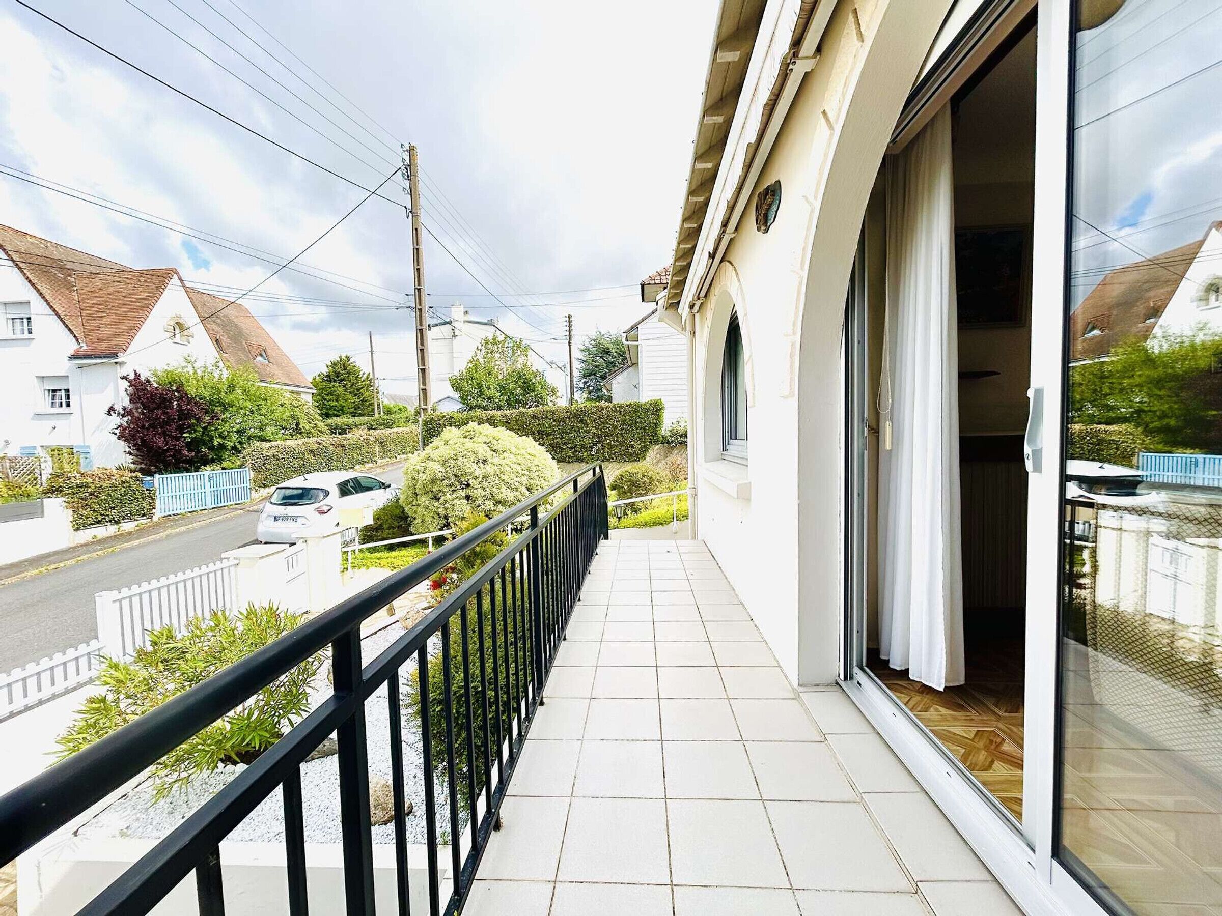 Un balcón residencial con barandilla negra, suelo de baldosas blancas y vista a una calle con casas y automóviles.