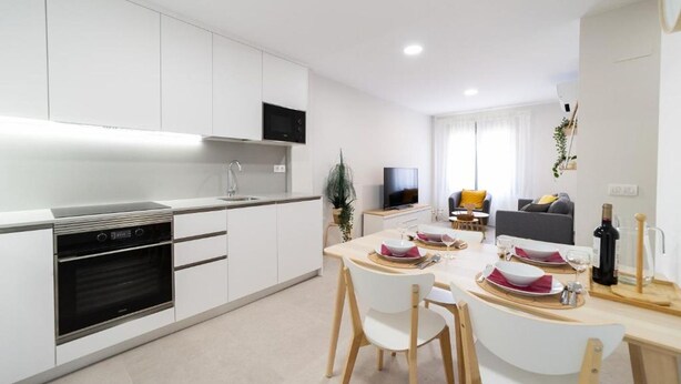 Una cocina moderna con armarios blancos, horno empotrado y comedor con mesa para dos.