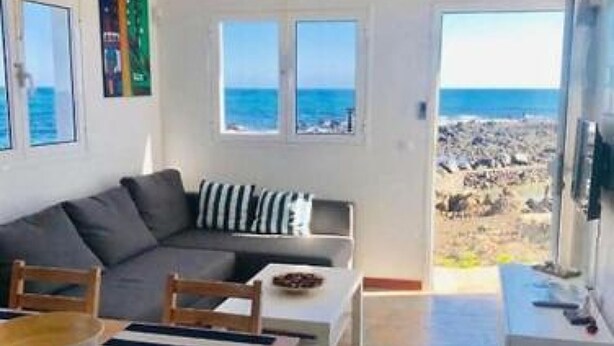 A modern living room with a sofa, dining table, and chairs. There is a view of the beach through a large window. - Fischer Home Roque del Este (Punta Mujeres)