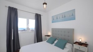A bedroom with a bed, two nightstands, a painting, a window with curtains, and a view of the sea. - Fischer Home Roque del Este (Punta Mujeres)