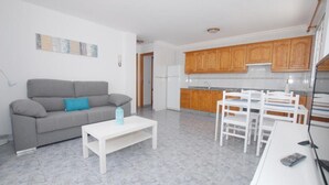 A compact living space with a grey sofa, a white coffee table, a dining table with chairs, wooden cabinets, and a white refrigerator. - Calma Suites La Garita B6 (Arrieta)