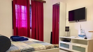A bedroom with a bed, red curtains, a TV mounted on the wall, a small shelf with a model ship, and a white cabinet.
