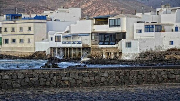 A coastal building with a stone wall in front, a rocky shoreline, and a mountainous backdrop.