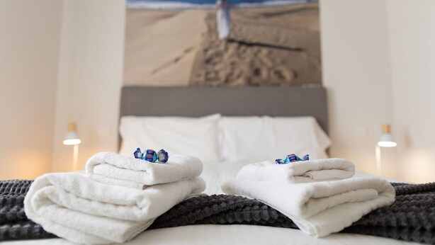 A neatly made bed with white towels and chocolates, a view of a beach with a person walking, and two bedside lamps.