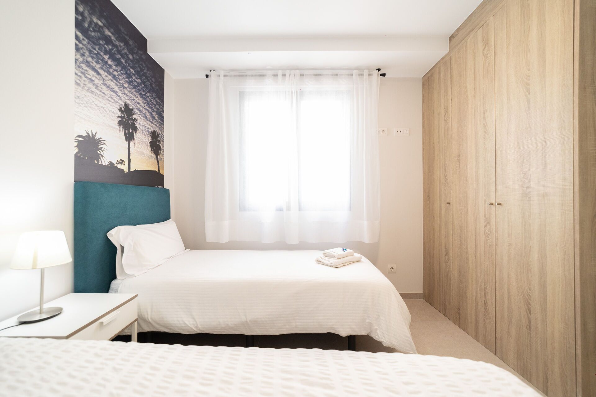 Une chambre à coucher comprenant un lit, une table de chevet, une lampe, une armoire et une fenêtre avec des rideaux légers.