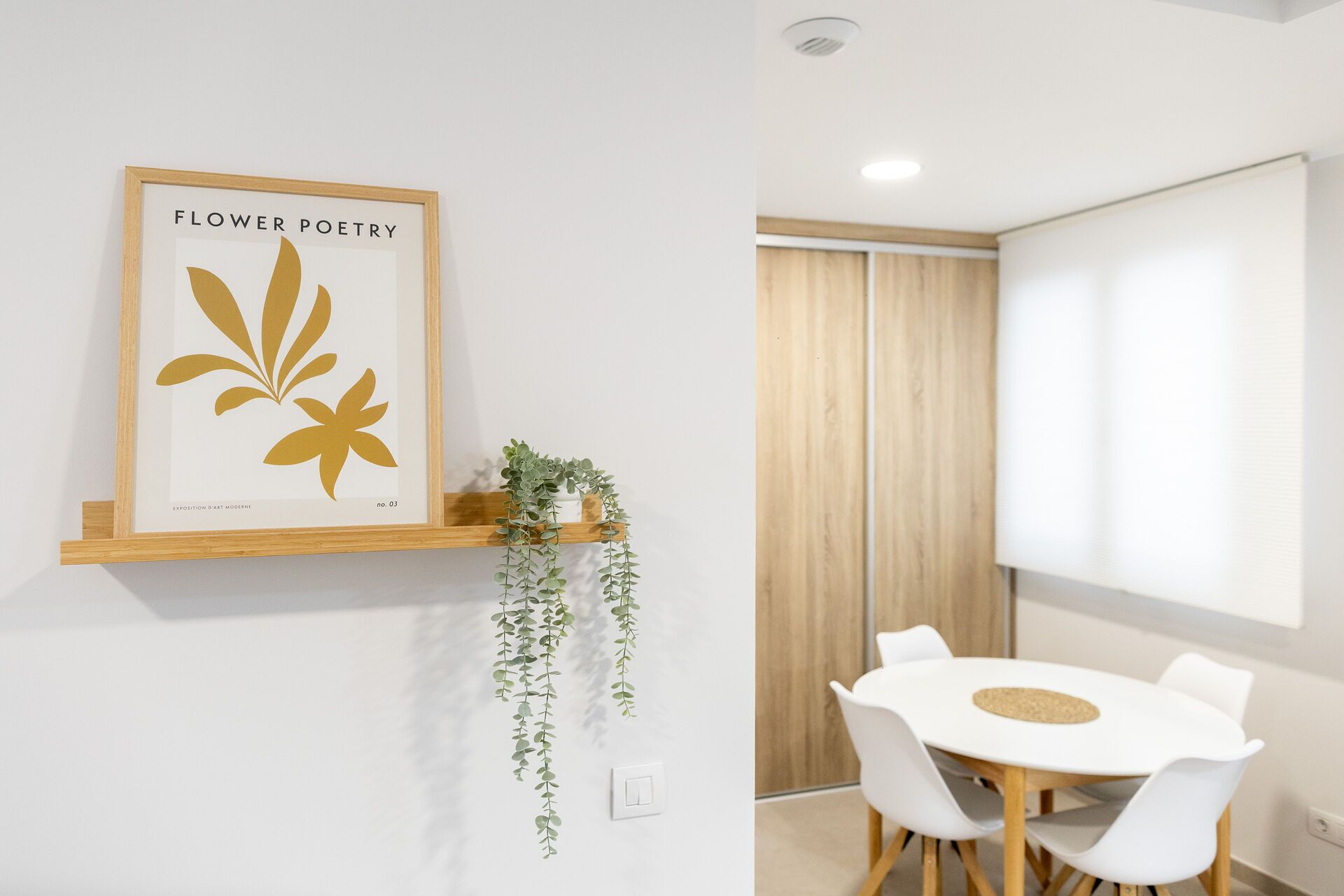 A modern room with a white table, chairs, a framed artwork titled 'Flower Poetry', and a hanging plant.