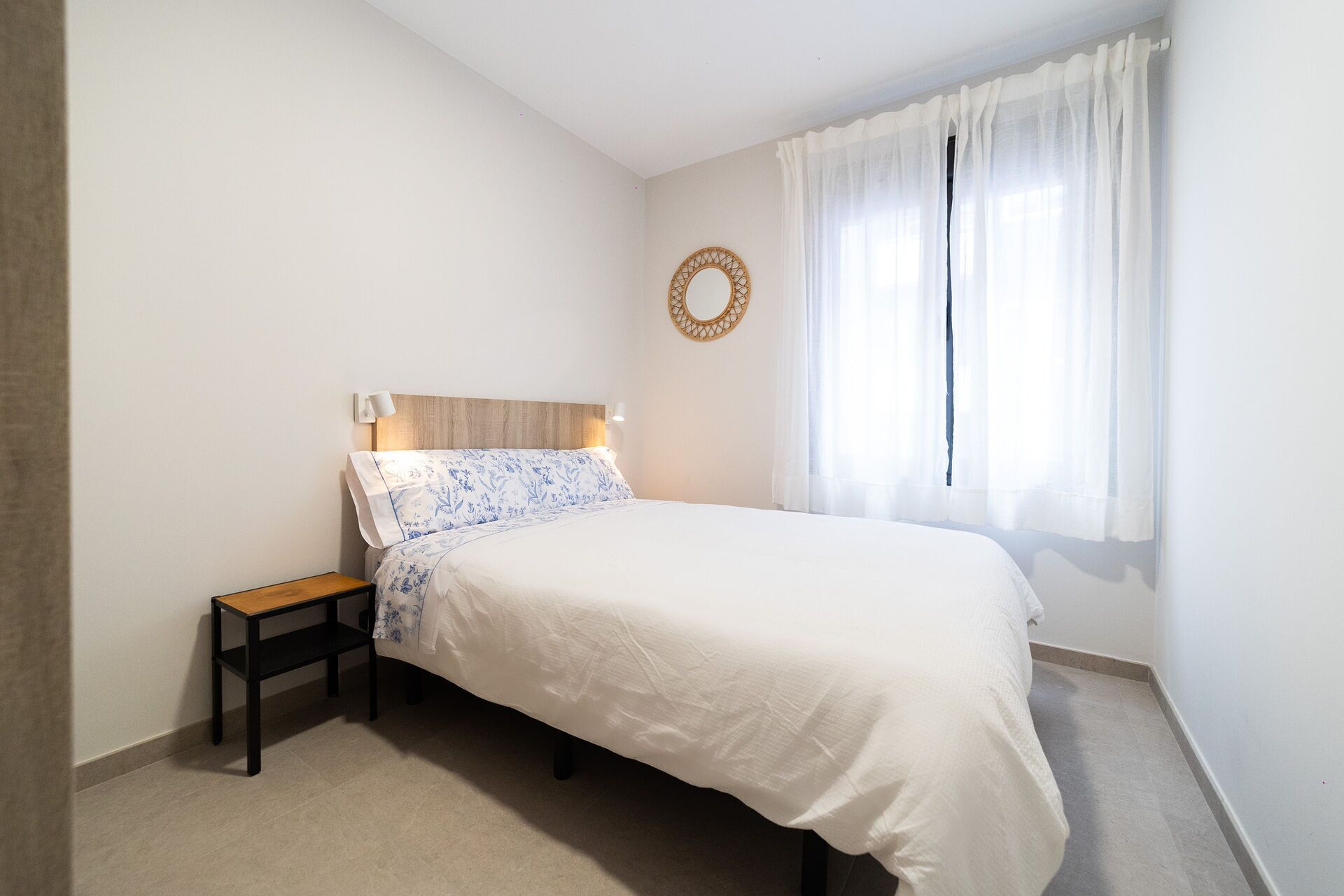 A bedroom with a bed, a nightstand, a wall-mounted mirror, and a window with sheer curtains.