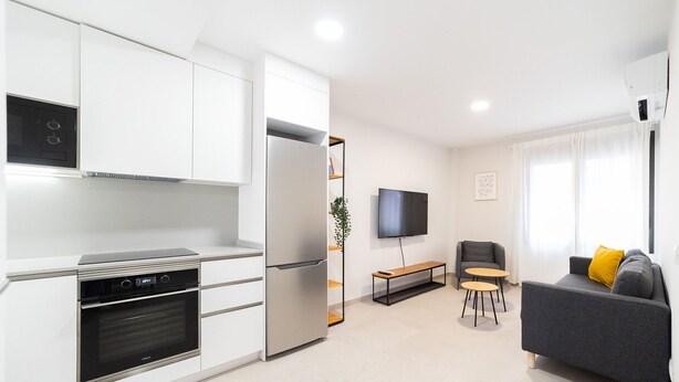 Una cocina moderna con armarios blancos, horno empotrado y refrigerador. Hay una zona de comedor con un banco y un televisor montado en la pared. Un sofá gris con un cojín amarillo está en la sala de estar.