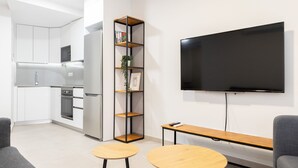 Una cocina moderna con armarios blancos y electrodomésticos de acero inoxidable, un televisor negro montado en la pared, y una pequeña zona de estar con dos mesas redondas y sofás grises.