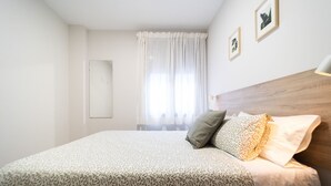 Un dormitorio con cama, cabecera, mesita de noche, espejo y ventana con cortinas.