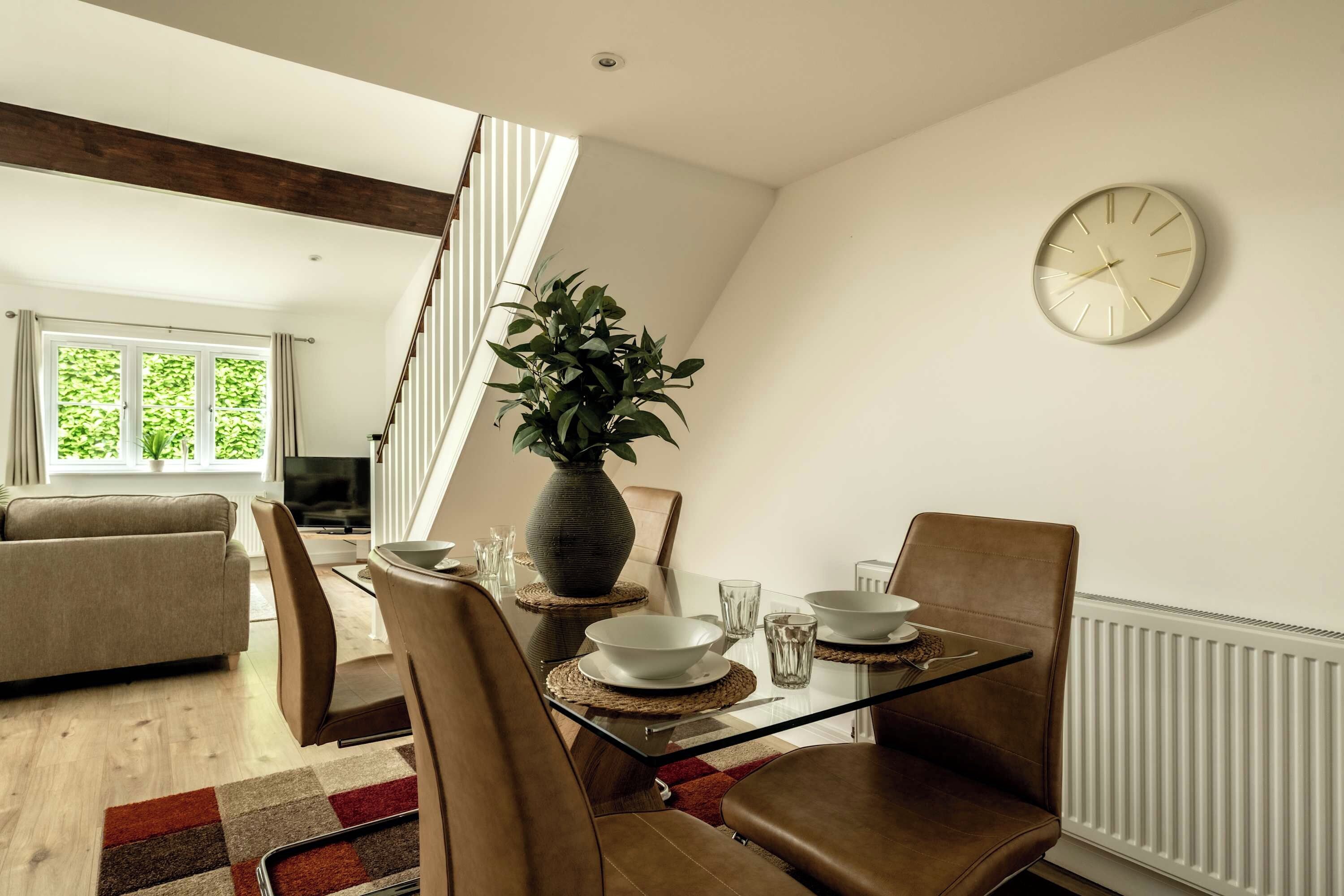 A dining area with a table set for two, a potted plant, and a clock on the wall.