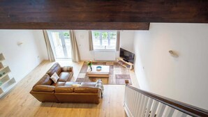 A spacious living room with a brown leather sofa, a coffee table, and a television. There is a staircase with a wooden railing leading to an upper floor. - York Cottage (South Cerney)