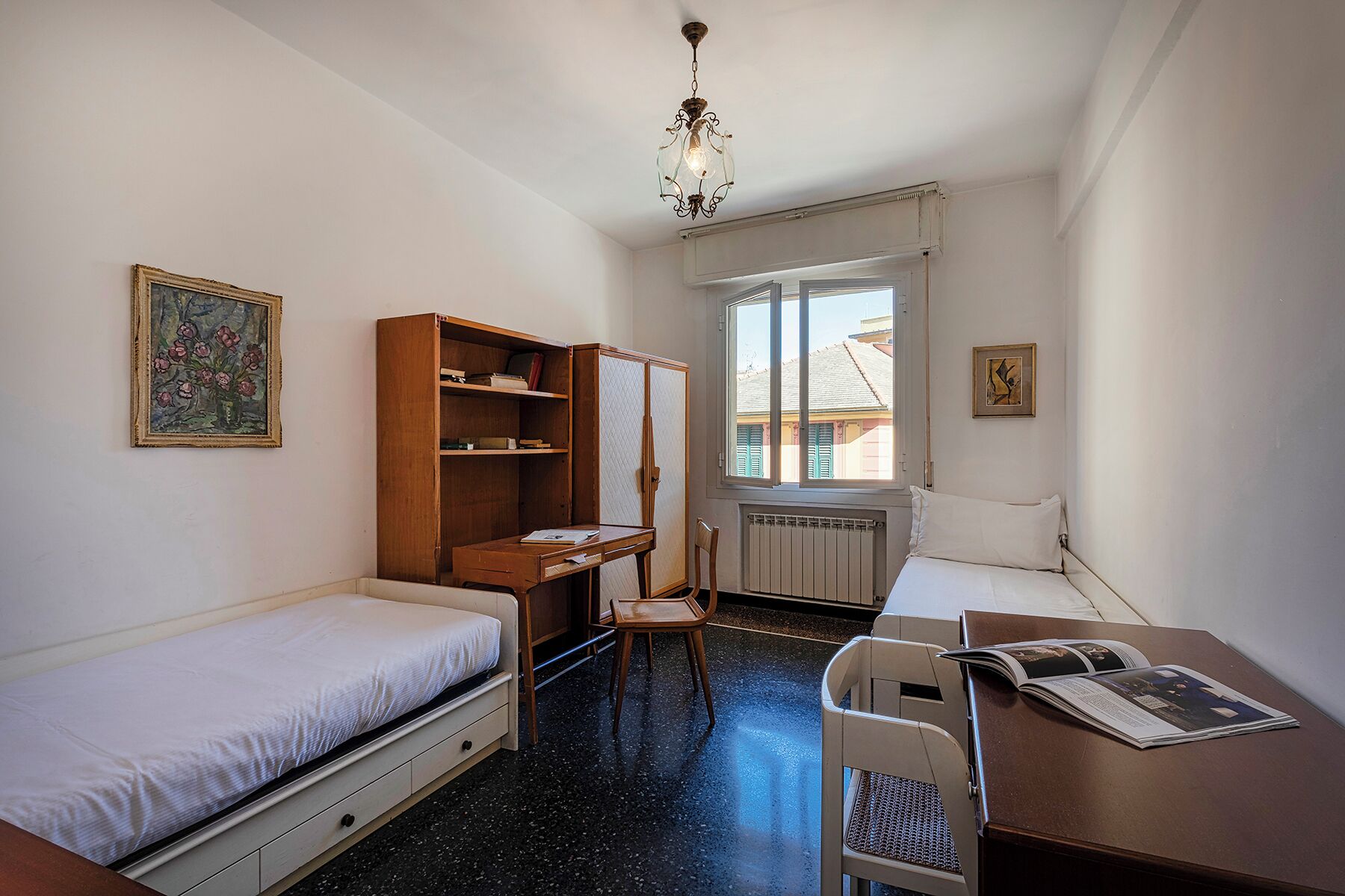Une petite pièce bien éclairée, avec un lit, un bureau, une chaise et un radiateur. Il y a une fenêtre qui donne sur les bâtiments à l’extérieur.