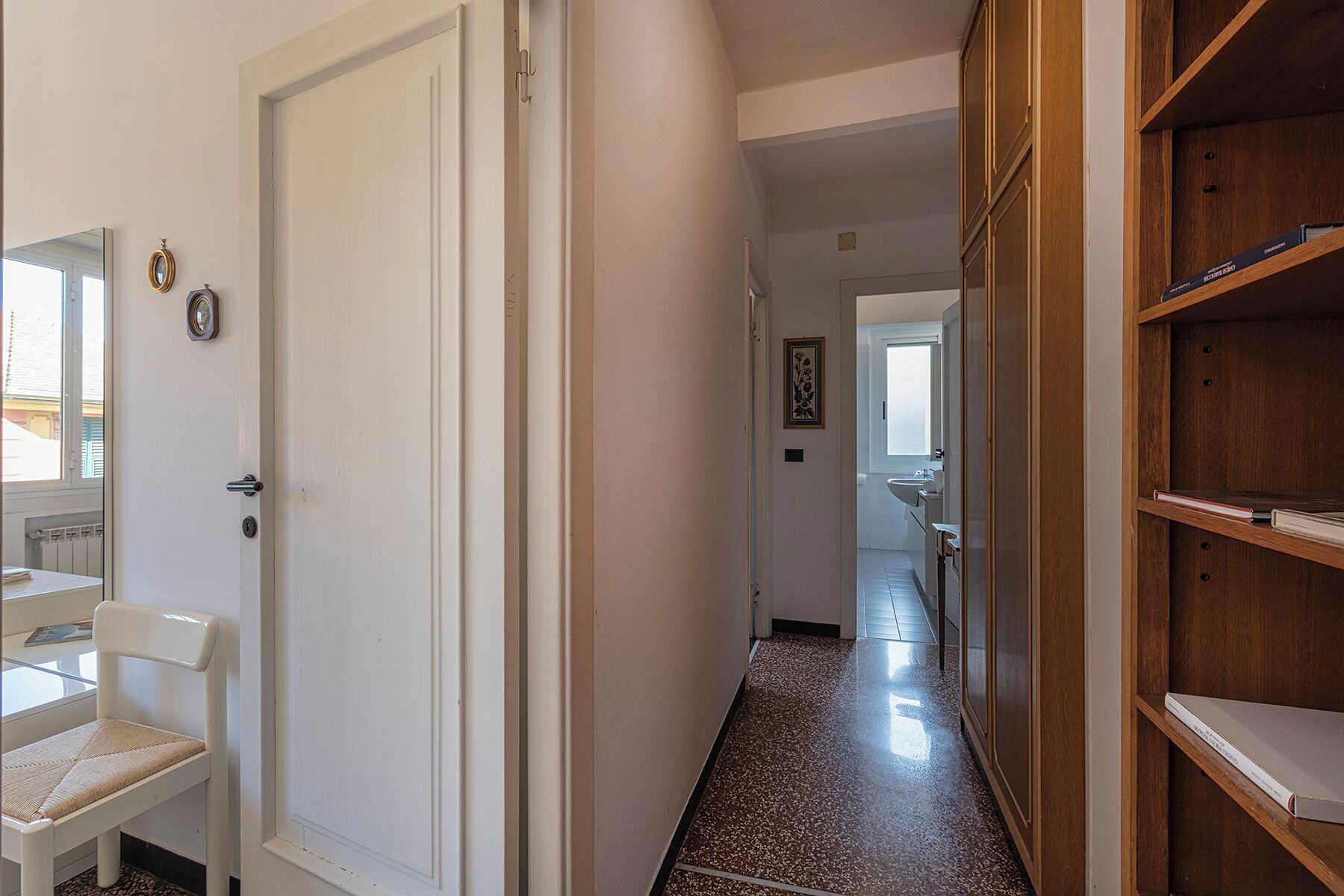 A narrow hallway with a white door on the left, a wooden bookshelf on the right, and a glimpse of a kitchen area in the distance.