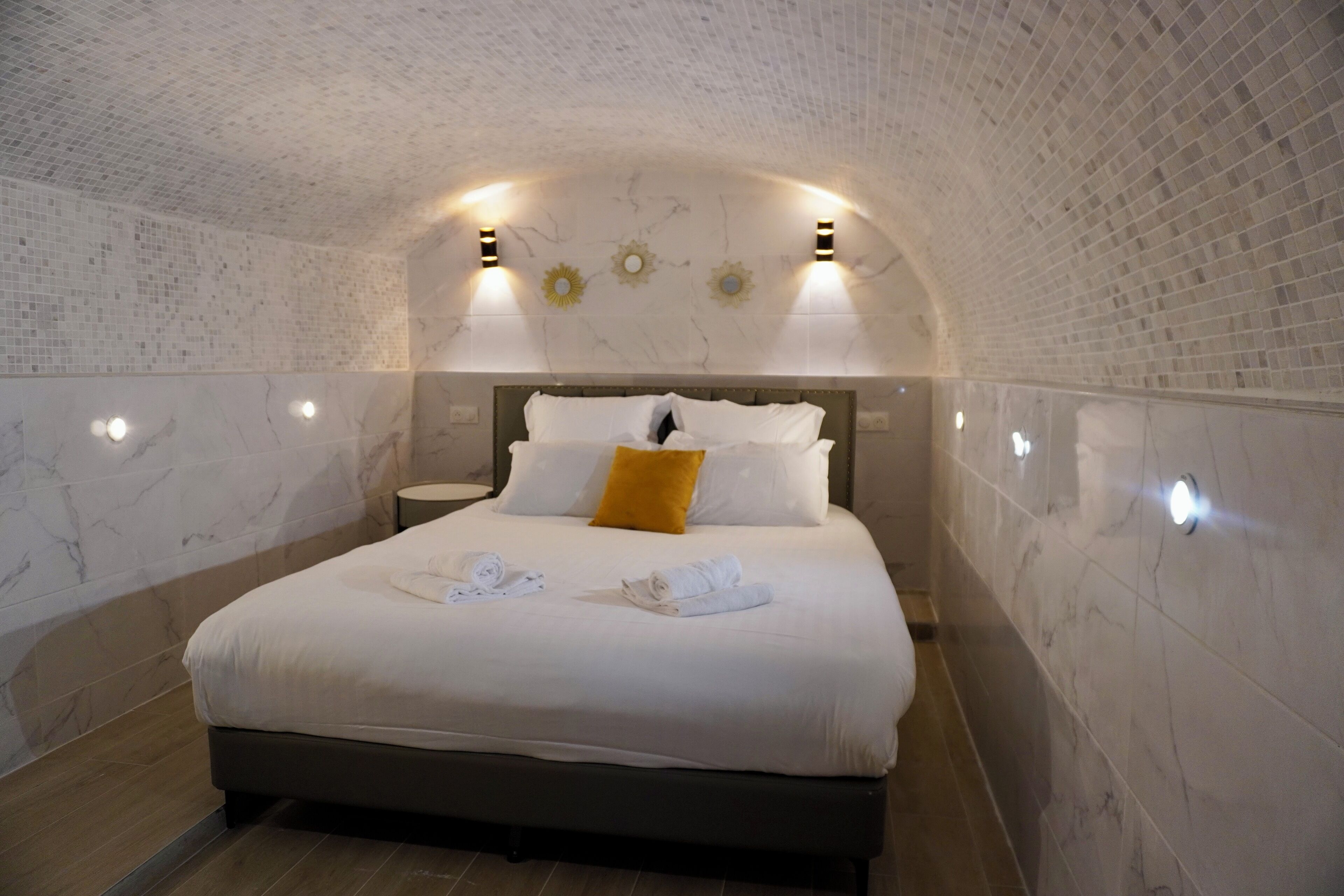 Quarto com cama, roupa de cama branca, almofada amarela e luminárias fixadas na parede.