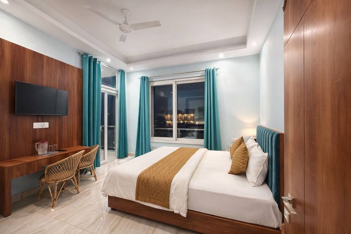 Quarto com cama, escrivaninha, cadeira, televisão e janela com cortinas.