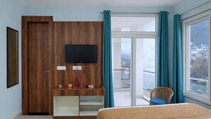 A hotel room with a bed, a TV mounted on a wooden panel, a balcony with a chair, and a view of the city. - Hotel DS valley Rishikesh (Narendranagar)