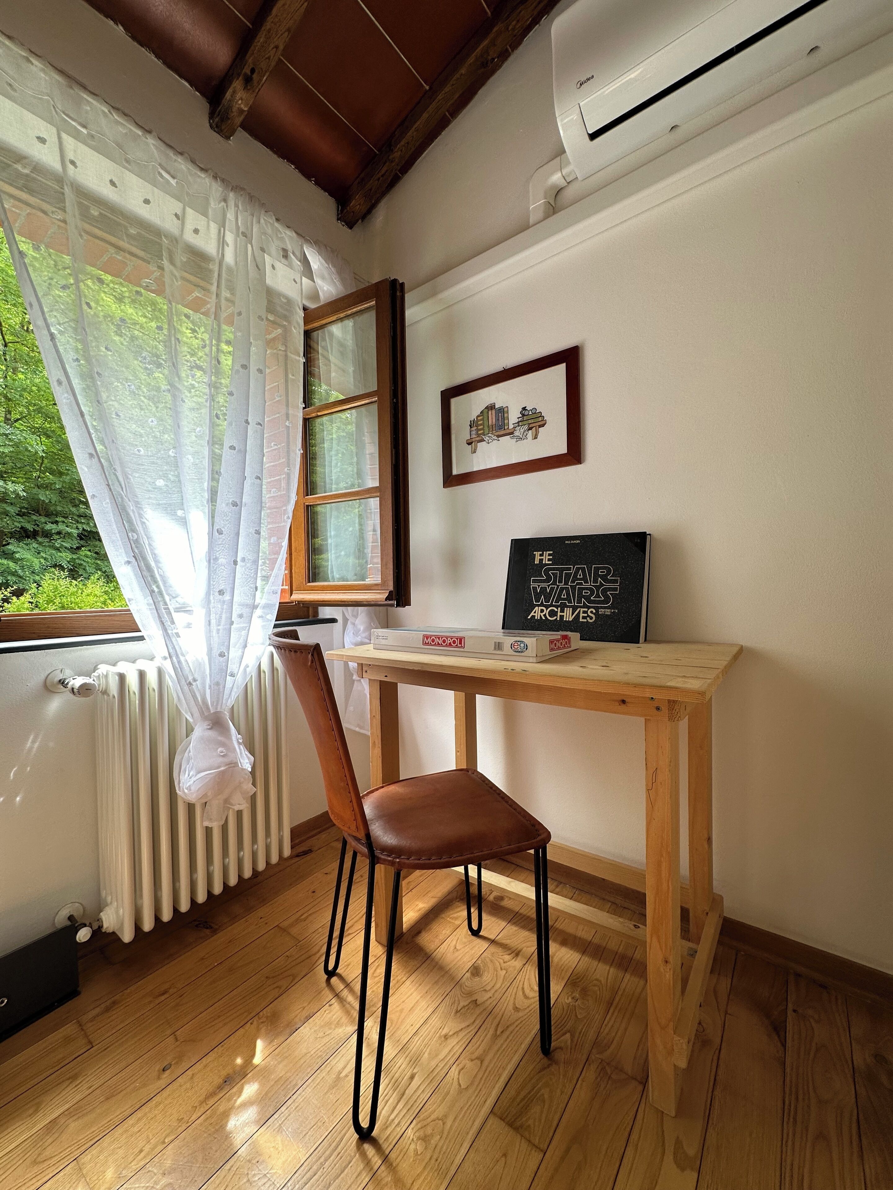 Una stanza con una scrivania in legno, una sedia, un termosifone, una finestra con tenda e un quadro appeso al muro.