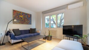 A modern living room with a sofa, a coffee table, a TV, and a large window with a view of buildings and trees.