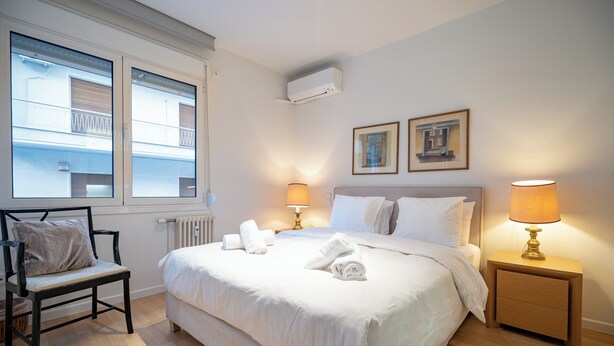 A hotel room with a large bed, a chair, a bedside table with a lamp, a window with blinds, and two framed pictures on the wall.