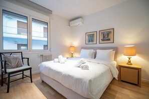 Habitación de hotel con una cama grande, una silla, una mesita de noche con lámpara, una ventana con persianas y dos cuadros en la pared.
