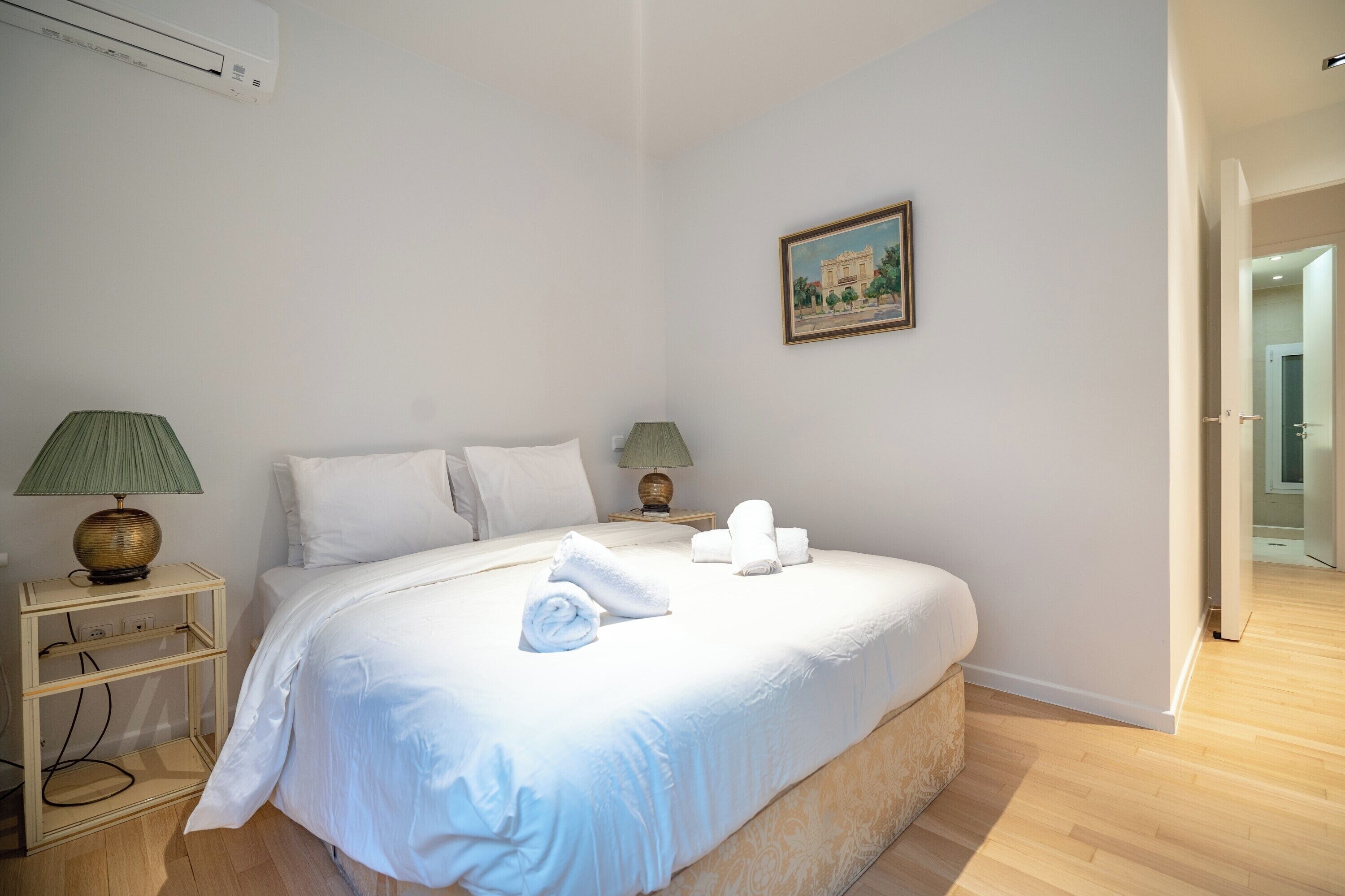 A bedroom with a bed, two bedside tables with lamps, a painting on the wall, and a door leading to another room.