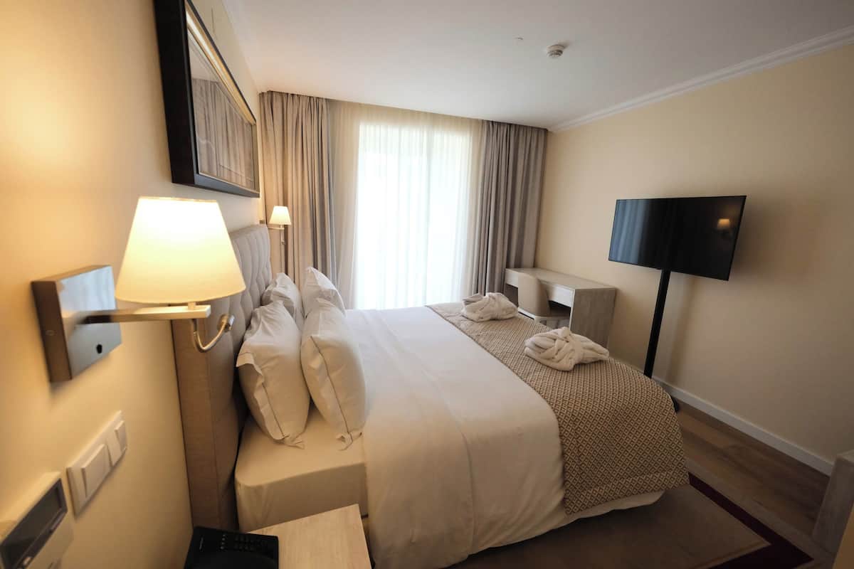 Ein Hotelzimmer mit einem großen Bett, einem Nachttisch, einem Fernseher und einem Fenster mit Vorhängen.