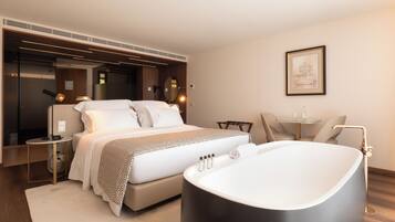 Una habitación de hotel moderna con una cama grande, una bañera, una mesita pequeña y una silla.