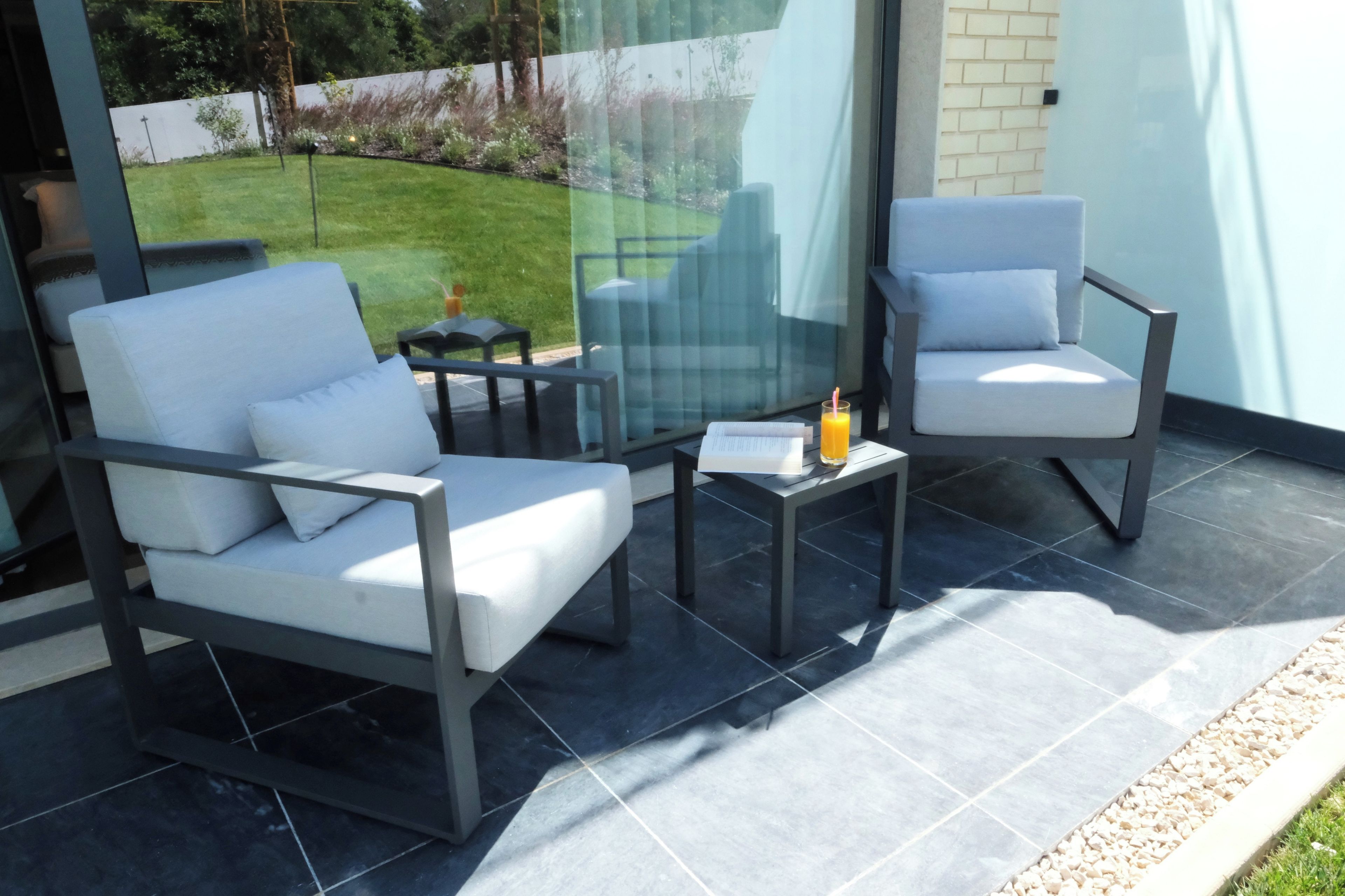 A modern outdoor seating area with two white cushioned chairs and a small table, featuring a glass partition and a view of a well-maintained lawn.