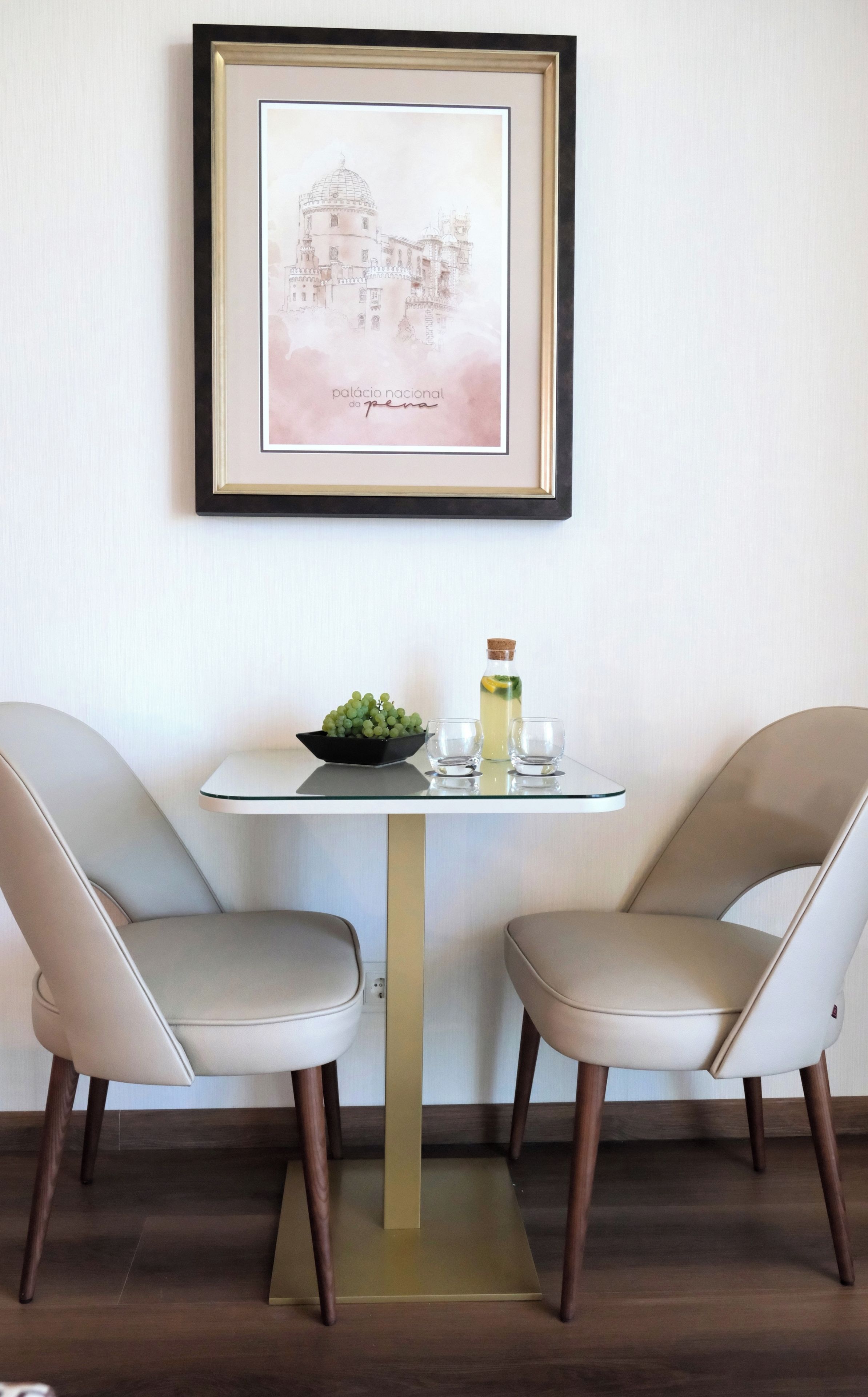 A dining area with two chairs, a table, a framed picture, a bowl of grapes, and a bottle of drink.