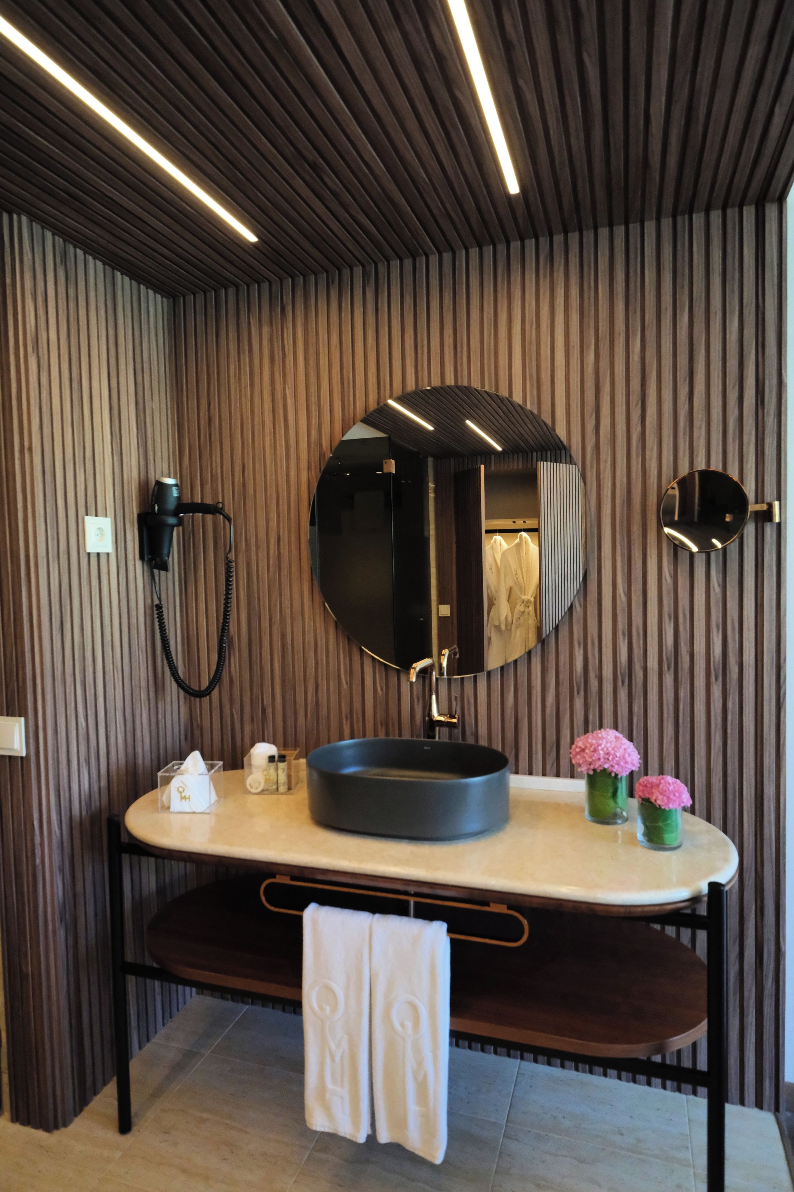 A modern bathroom with a wooden panel wall, a round mirror, a black sink, and a towel rack.