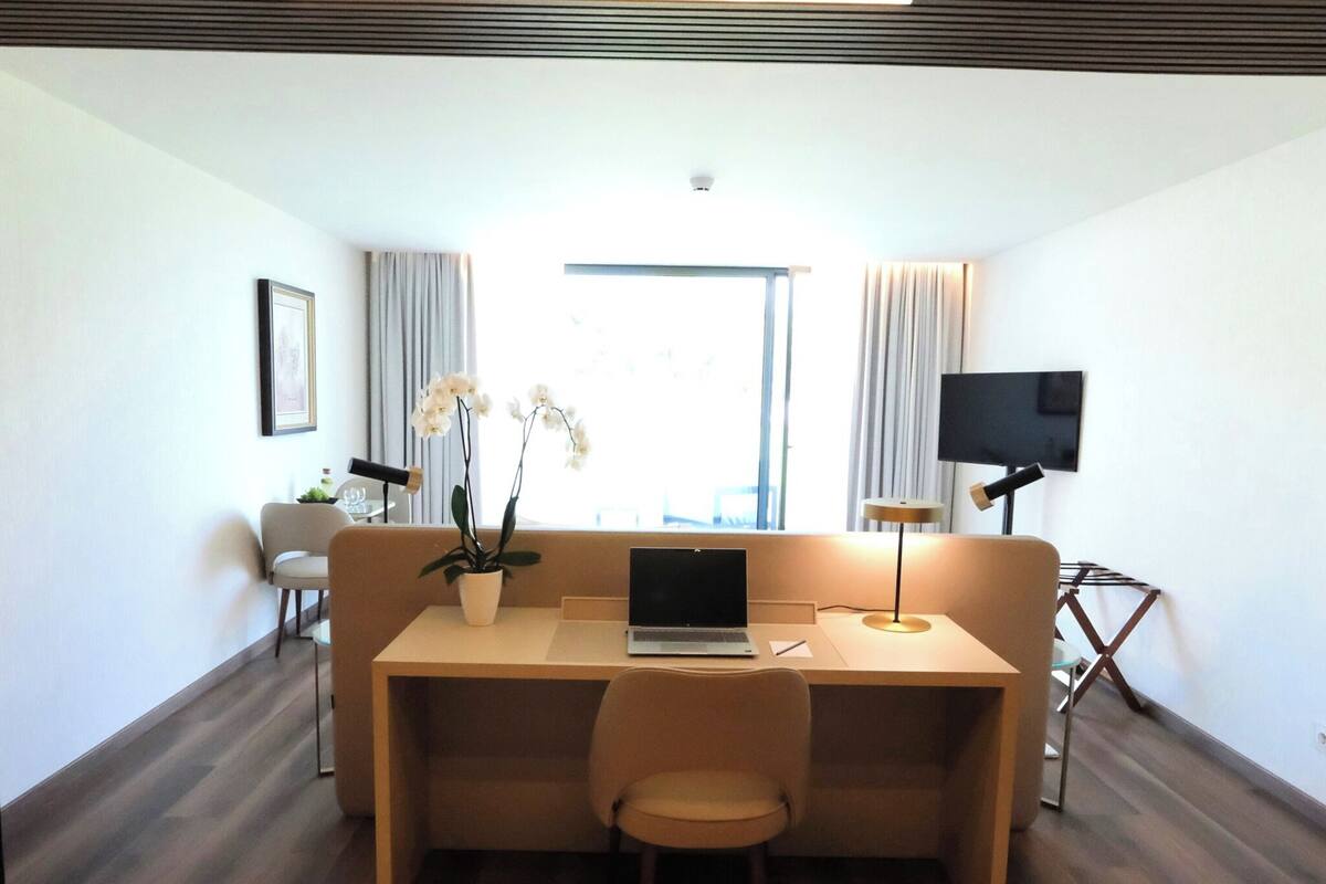 Ein modernes Zimmer mit Holzboden, einer Couch, einem Schreibtisch mit Laptop, einem Fernseher und einem Fenster mit Vorhängen.