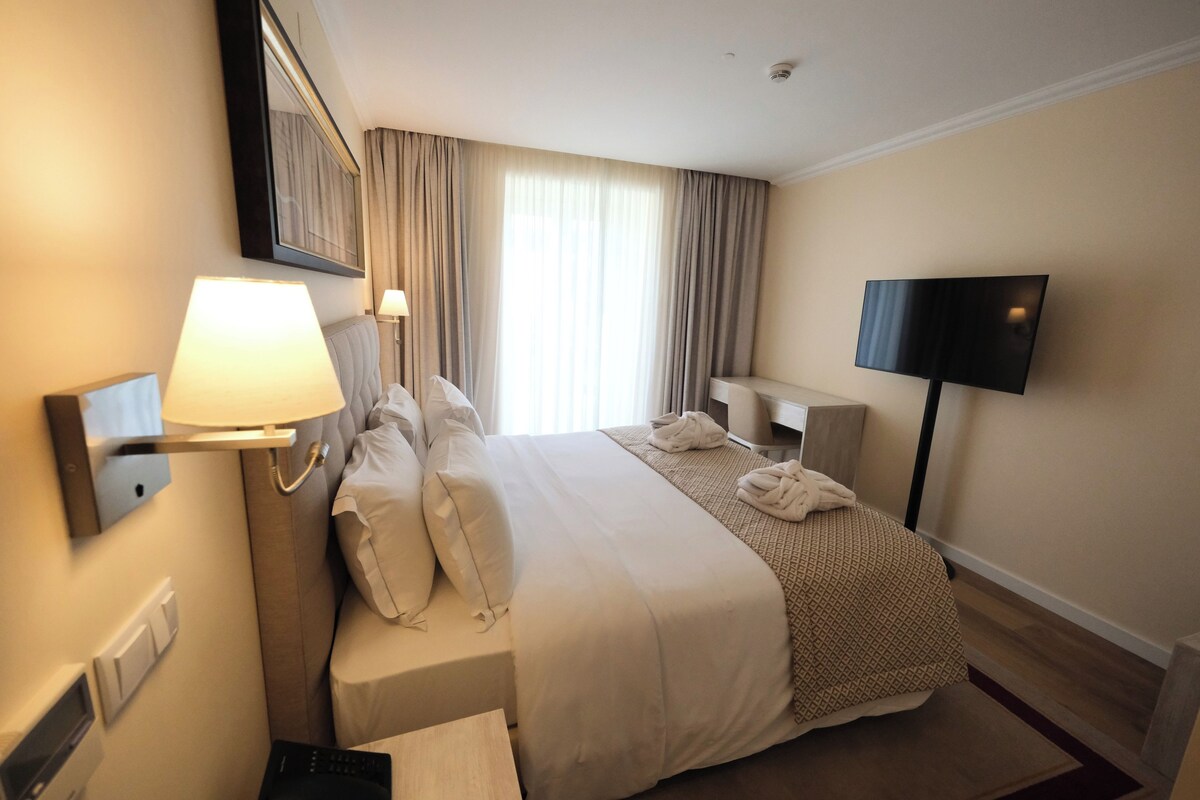 A hotel room with a large bed, a bedside table, a TV, and a window with curtains.