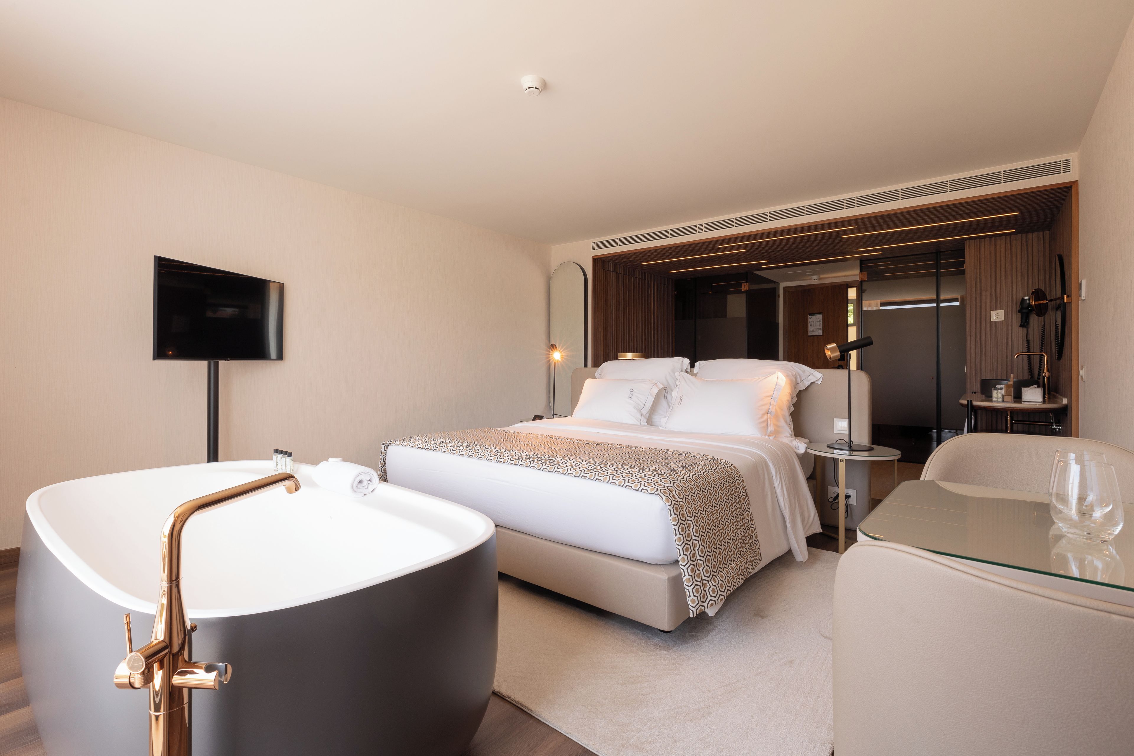 Una habitación de hotel moderna con una bañera grande, una cama con cubrecama estampado, una mesa de centro de vidrio y un televisor de pantalla plana montado en la pared.