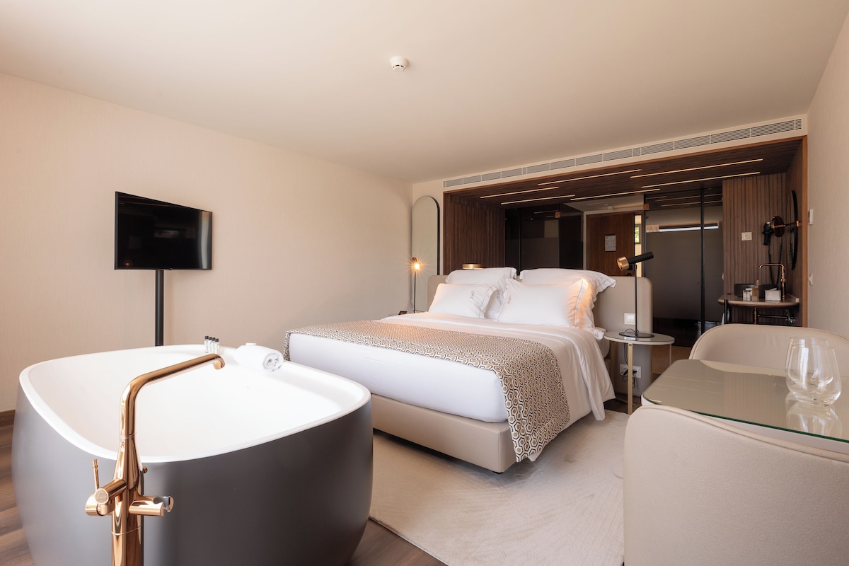 A modern hotel room with a large bathtub, a bed with a patterned bedspread, a glass coffee table, and a flat-screen TV mounted on the wall.