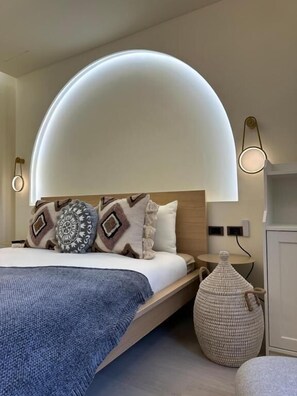 Un dormitorio moderno con una lámpara de techo curva, una cama con almohadillas estampadas, una mesita de noche y un otoman tejido.