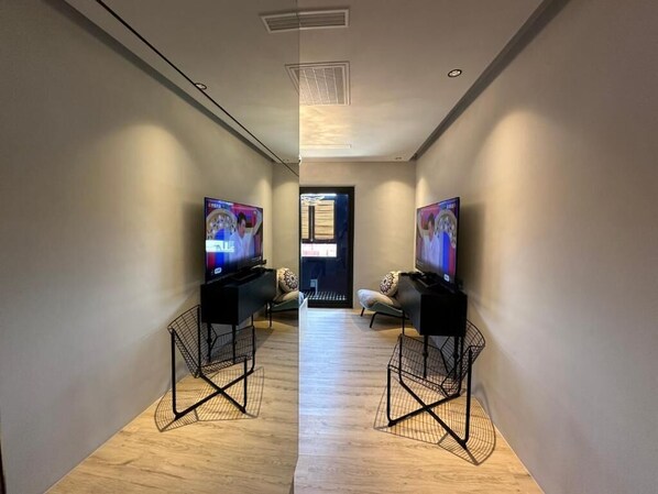 Un pasillo con dos televisores sobre soportes negros, piso de madera y una puerta al final.