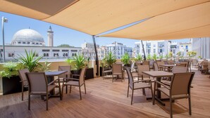 Een terras met tafels en stoelen, plantenpotten en uitzicht op een moskee met koepel en minaret.