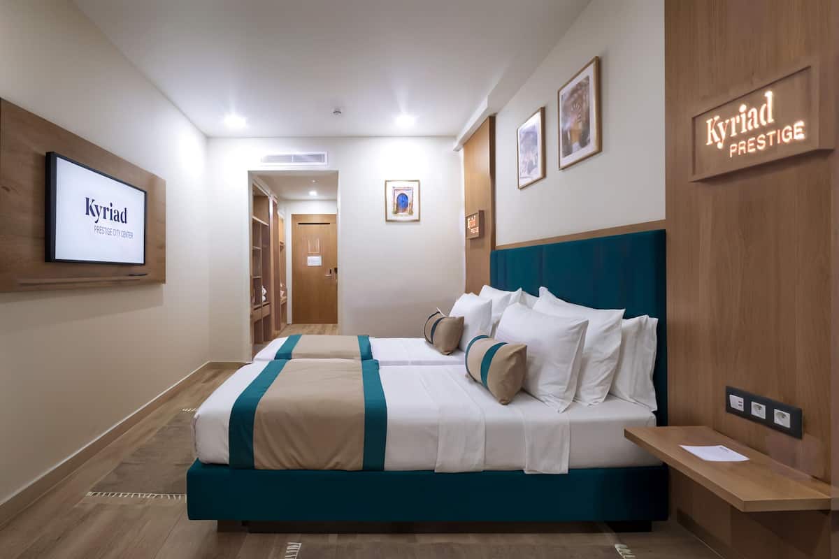 Ein Hotelzimmer mit einem großen Bett, einem Schreibtisch und einem Kleiderschrank. An den Wänden befinden sich Bilder in Rahmen und ein Schild mit der Aufschrift 'Kyriad Prestige'.