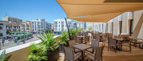 An outdoor terrace with tables and chairs, potted plants, and a view of the cityscape.