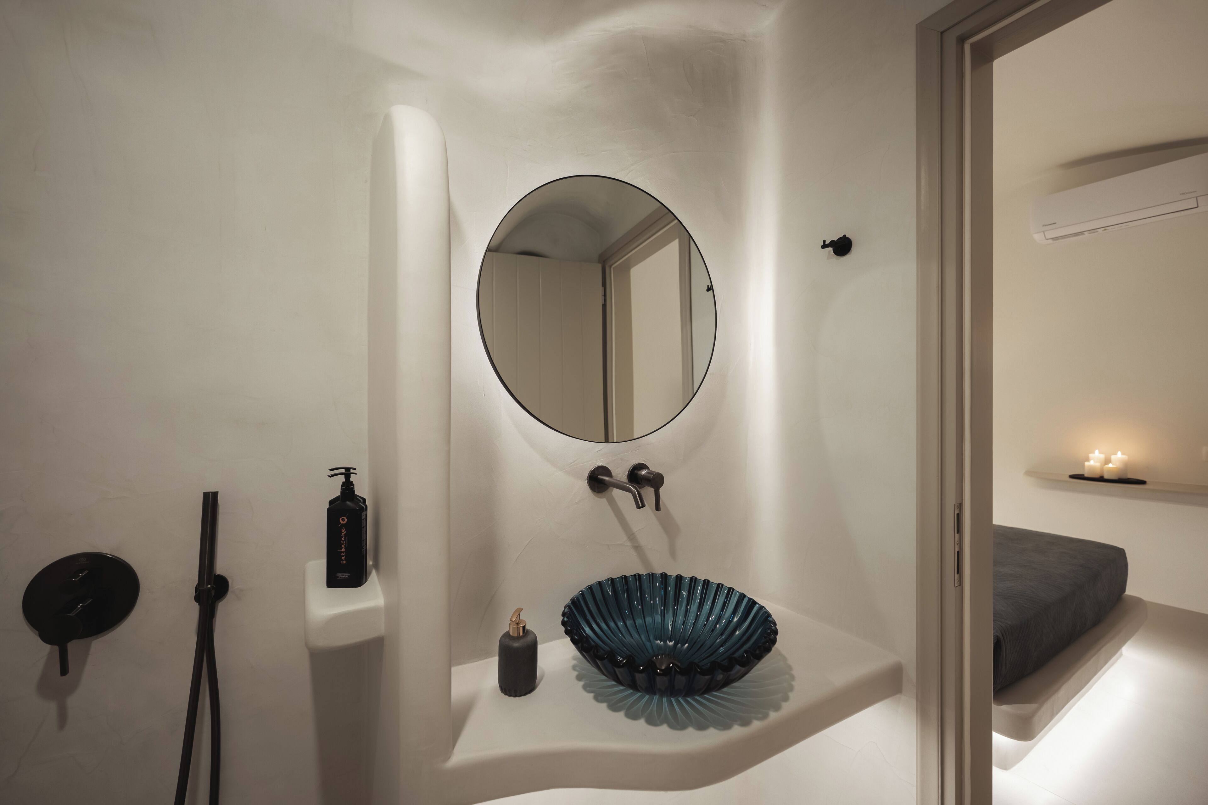 Un baño con espejo redondo, lavamanos con cuenca azul, ducha y mesita de noche con vela.